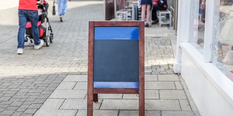 Ein unbeschrifteter Aufsteller aus Holz steht vor einem Geschäft in einer Fußgängerzone. Im Hintergrund ist die Außenbestuhlung eines Cafés. Neben dem Aufsteller laufen Passanten. 